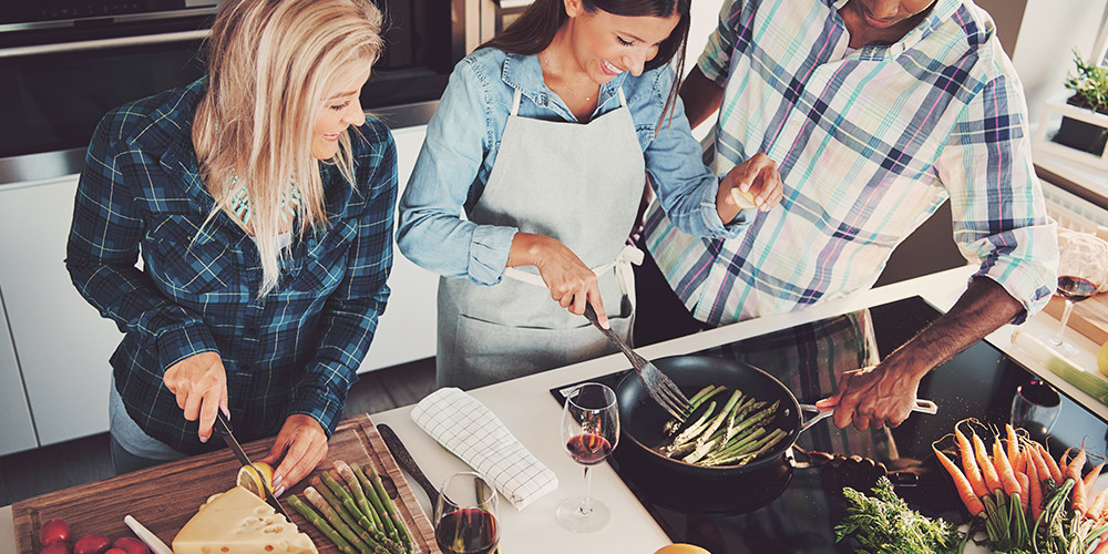 Drie mensen koken samen in een keuken, waarbij ze groenten bereiden op een kookplaat.