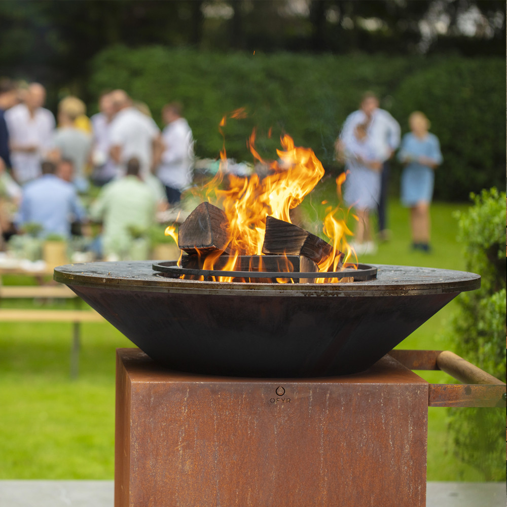 Vuurkorf met brandend hout in een tuin, met mensen op de achtergrond.