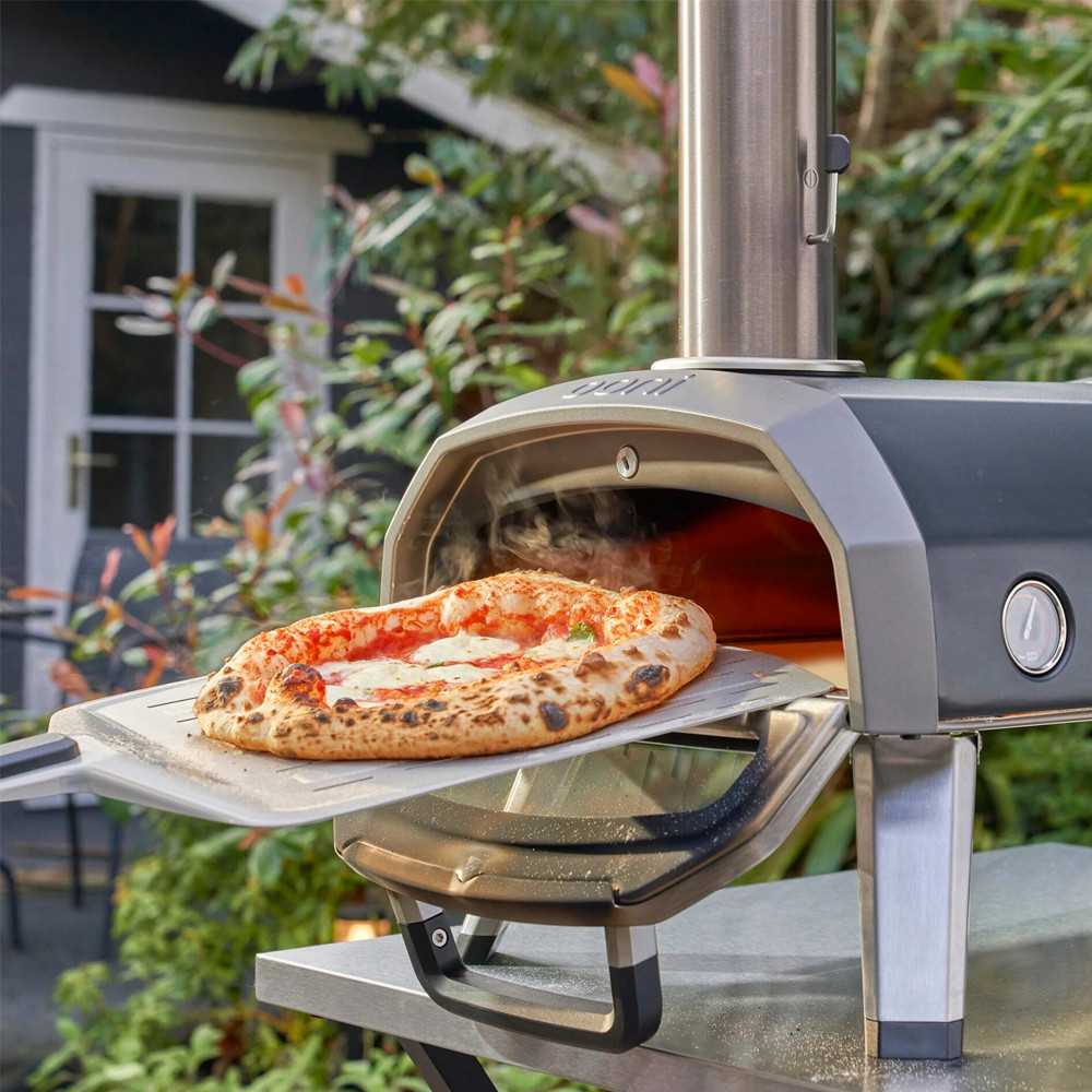 Pizza wordt uit een roestvrijstalen houtgestookte oven gehaald in een tuinomgeving.