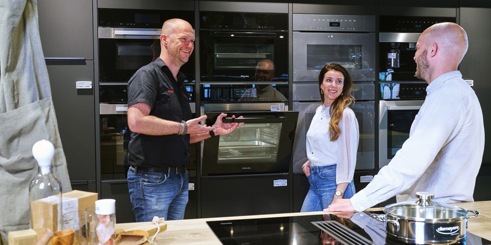 Drie mensen staan lachend in een keukenshowroom en bekijken inbouwapparatuur.