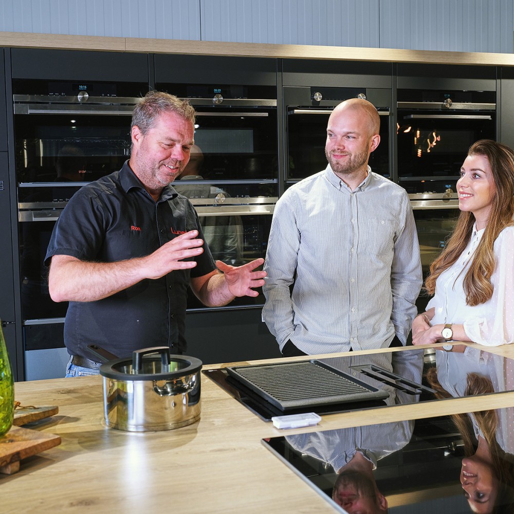 Drie mensen staan in een moderne keuken, pratend naast een kookplaat met een pan en grillplaat.