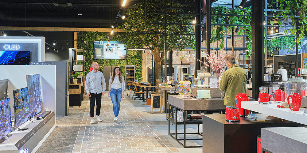 Een moderne winkel met elektronica en keukenapparatuur, groene planten en klanten die rondlopen.
