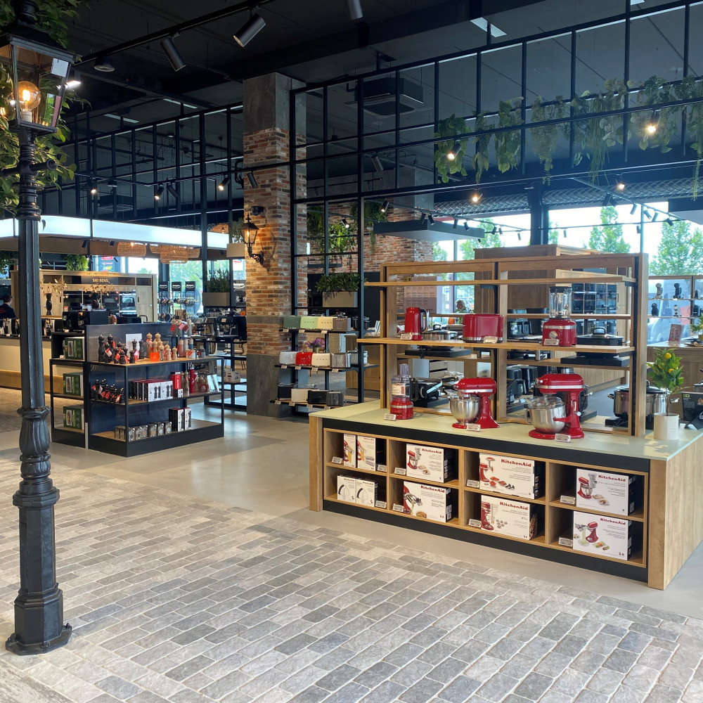 Winkelinterieur met keukenapparatuur op houten rekken, bakstenen pilaren en een glazen wand.