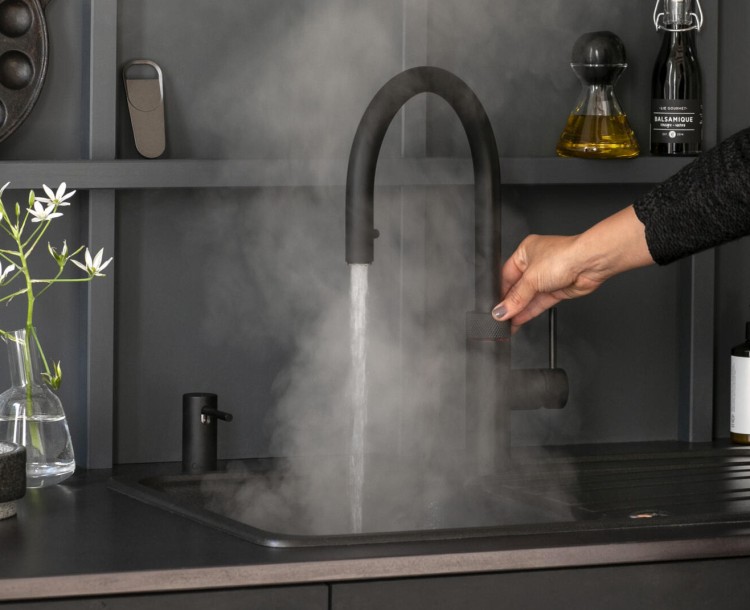 Een persoon bedient een keukenkraan waaruit stoom uitkomt, naast een vaas met witte bloemen.