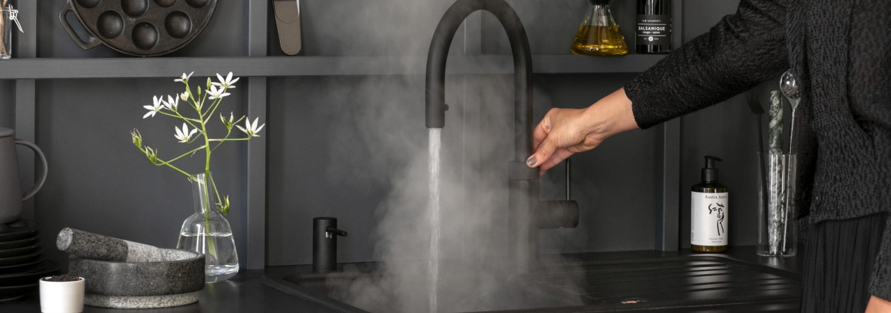 Een persoon bedient een keukenkraan waaruit stoom uitkomt, naast een vaas met witte bloemen.