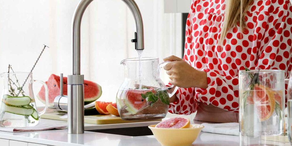 Persoon in rode stippenblouse vult een kan met fruitig water bij een moderne keukenkraan.