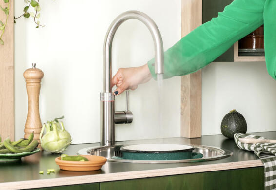Een persoon bedient een moderne keukenkraan, omgeven door groenten en keukenspullen.