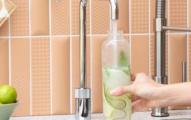 Hand vult een glazen fles met water onder een kraan in een keuken met beige tegels.