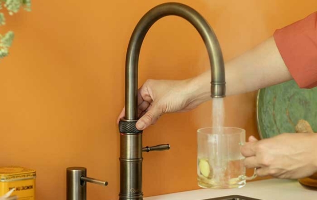 Een persoon vult een glas met heet water uit een moderne keukenmengkraan tegen een oranje muur.