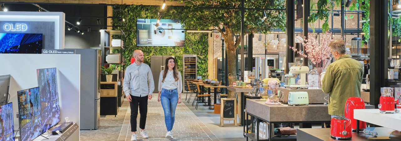 Een paar mensen in een moderne winkel met elektronica en keukenspullen, omgeven door groen.