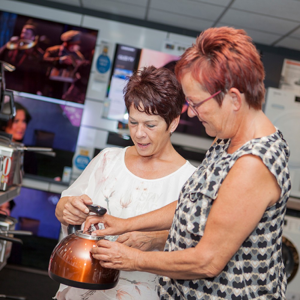 Twee vrouwen bekijken een object in een winkel vol met elektronische apparaten.