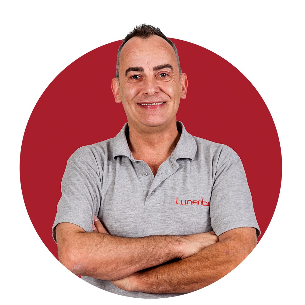 Een man in een grijs poloshirt tegen een rode achtergrond, met gekruiste armen en glimlachend.