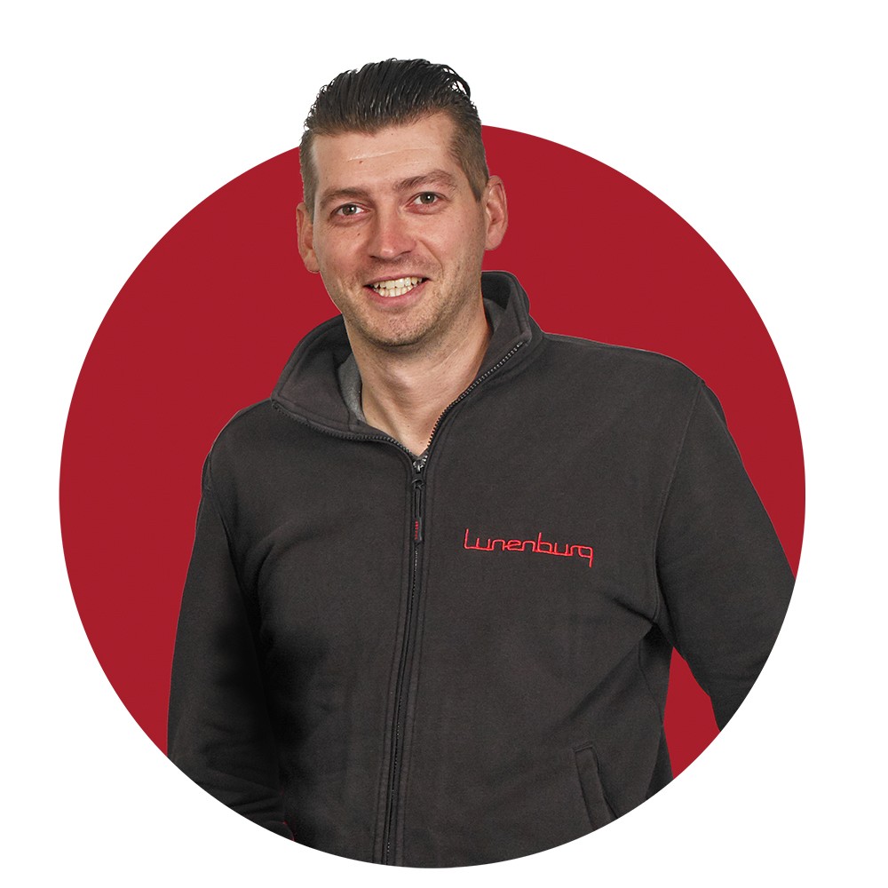 Een man in een zwarte trui met rood logo, op een rode cirkel achtergrond.