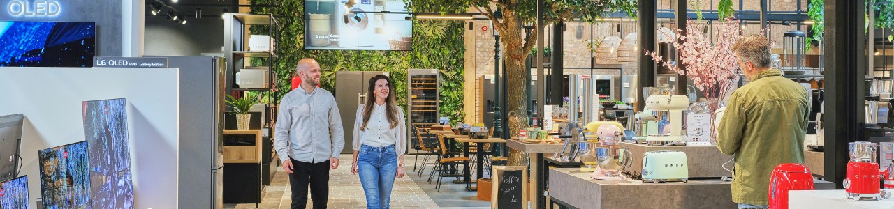 Mensen lopen in een moderne winkel met tv's, keukenapparatuur en planten.