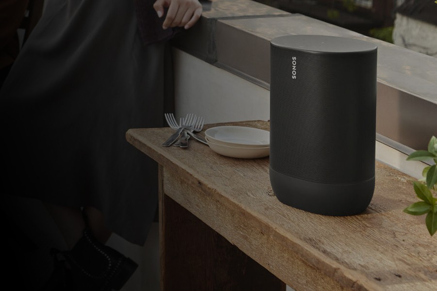 Zwarte luidspreker op een houten tafel buiten, met een bord, bestek en een plant naast de luidspreker.